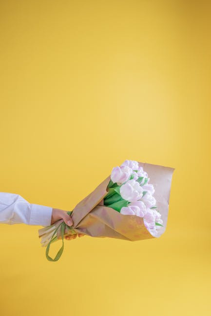A delivery person wearing a red cap and red jacket holds a colorful gift box with a geometric pattern, possibly containing a floral arrangement or gift. The individual is standing outside near a white
