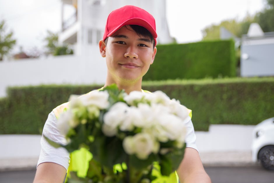 A delivery person wearing a red cap and a high-visibility vest is holding out a bouquet of fresh white flowers toward the camera. The bouquet features tightly packed white petals with green stems and 