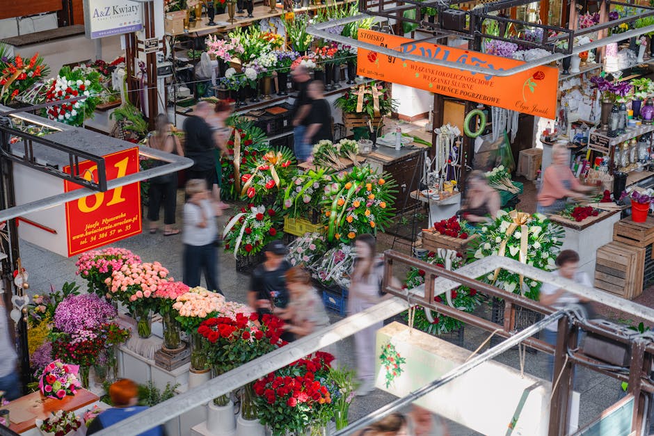 Indoor flower shop bustling with customers, featuring various floral arrangements and bouquets. Visible are vibrant pink, red, purple, and white flowers arranged in vases and baskets, with some wrappe