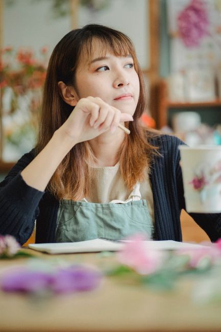 A close-up of a woman with shoulder-length auburn hair and bangs, holding a paintbrush near her mouth while observing a floral arrangement in front of her. She is wearing a cream-colored top under a d
