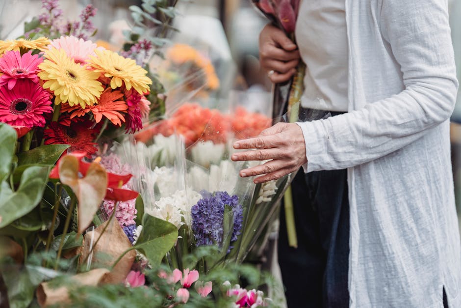 A person wearing a light grey cardigan and dark trousers is browsing a flower stall, examining a bouquet of fresh flowers wrapped in transparent plastic. The display includes vibrant pink, yellow, pur